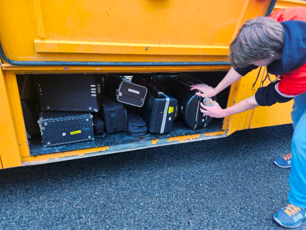  A Game of Tetris: Kamren Meloy stuffing the bus with his trombone case. When the Cashmere High School football team qualified for the state championship game, a large part of the excitement didn’t just come from the fans who’d get to watch the game, or from the team who’d get to play, but from the pep band. They would be performing at one of the most important games in CHS sports history, and as far as anyone was aware, the band might never get the opportunity to do anything like this ever again. Before the band could leave the high school, they went through their first obstacle: loading the bus. Band director Mr. Chalmers has often compared doing so to “playing Tetris,” and he’s not wrong. With several dozen band members all trying to put their stands and instruments into one school bus luggage box, it can be quite a pain to fit everything. For example, Kamren Meloy, pictured above, had a difficult time putting his large trombone case into the cramped heap of cases and gigbags.

